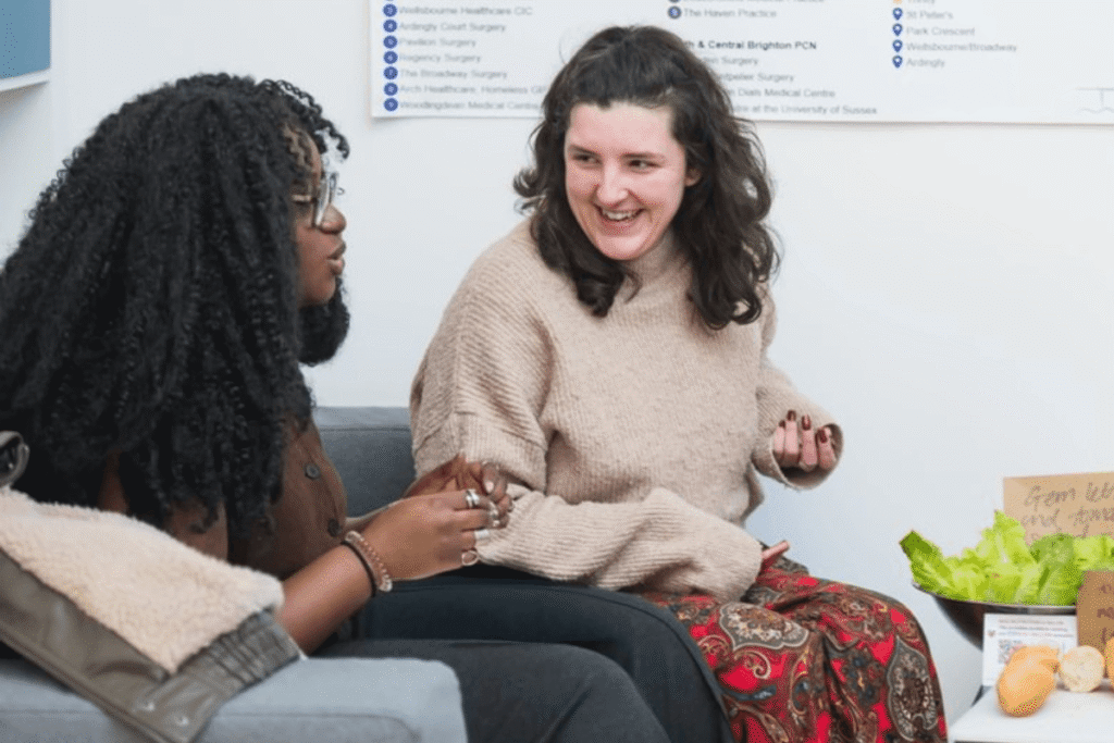 Two female Brighton and Hove GP Federation staff members sitting on a grey couch, engaged in a cheerful conversation. The woman on the left, with long dark textured hair and glasses, is speaking and gesturing with her hands. The woman on the right, wearing a beige sweater, is smiling and laughing warmly. In the background, a poster listing local surgeries is partially visible on the wall, and a small table with fresh vegetables and a bowl of lettuce sits to the right.