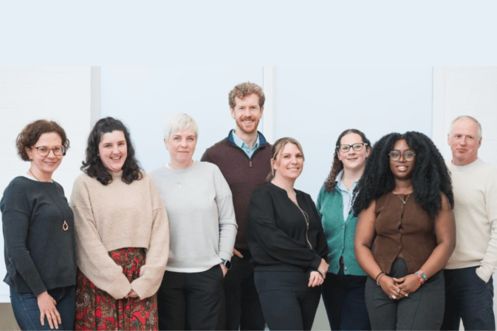 A group photograph of eight Brighton and Hove GP Federation staff members standing together indoors. They are dressed in casual and smart-casual clothing, smiling warmly at the camera against a brightly lit, plain background.